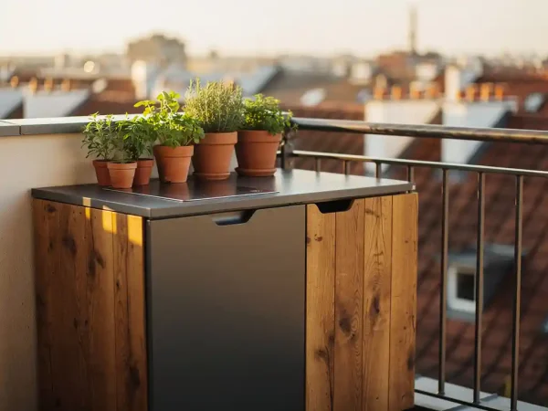 Compacte buitenkeuken van staal en eikenhout op stadsbalkon met kruidenplanten in terracotta potten, gouden middaglicht.