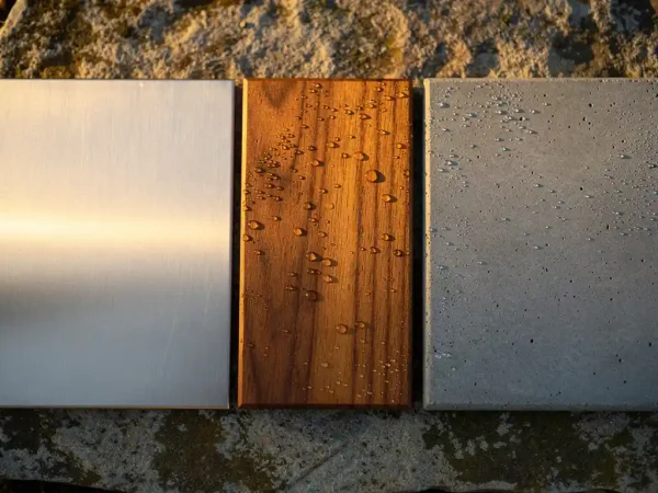 Brushed roestvrijstalen plaat, teakhouten plank en betonnen tegel op verweerd steen in warm ochtendlicht.