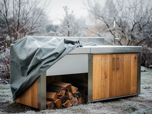 Buitenkeuken van staal en hout met lichte rijp, halfafgedekt met weerbestendig hoes, stapel brandhout ertegenaan, kale wintertuintakken op de achtergrond.