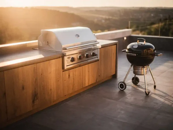 Ingebouwde roestvrijstalen grill in buitenkeuken met houten kasten en betonnen werkblad, naast vrijstaande houtskoolbarbecue op zonnig terras.