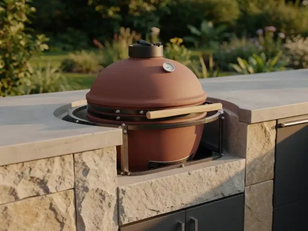 Keramische kamado-grill ingebouwd in natuurstenen buitenkeukenblad, omgeven door stalen frame en beton, weelderige tuin op de achtergrond.